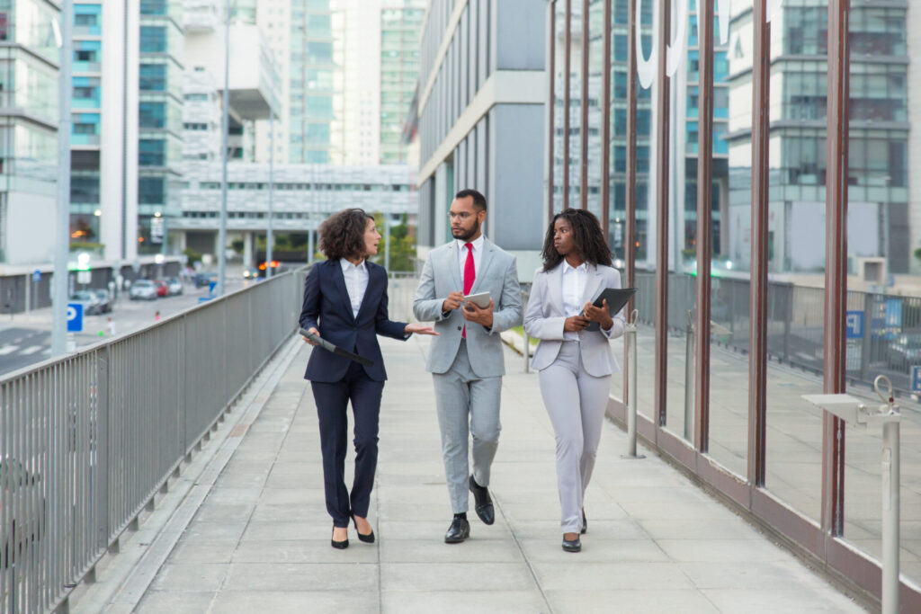 A group of three diverse business professionals walking and discussing on a city street, with modern office buildings in the background.